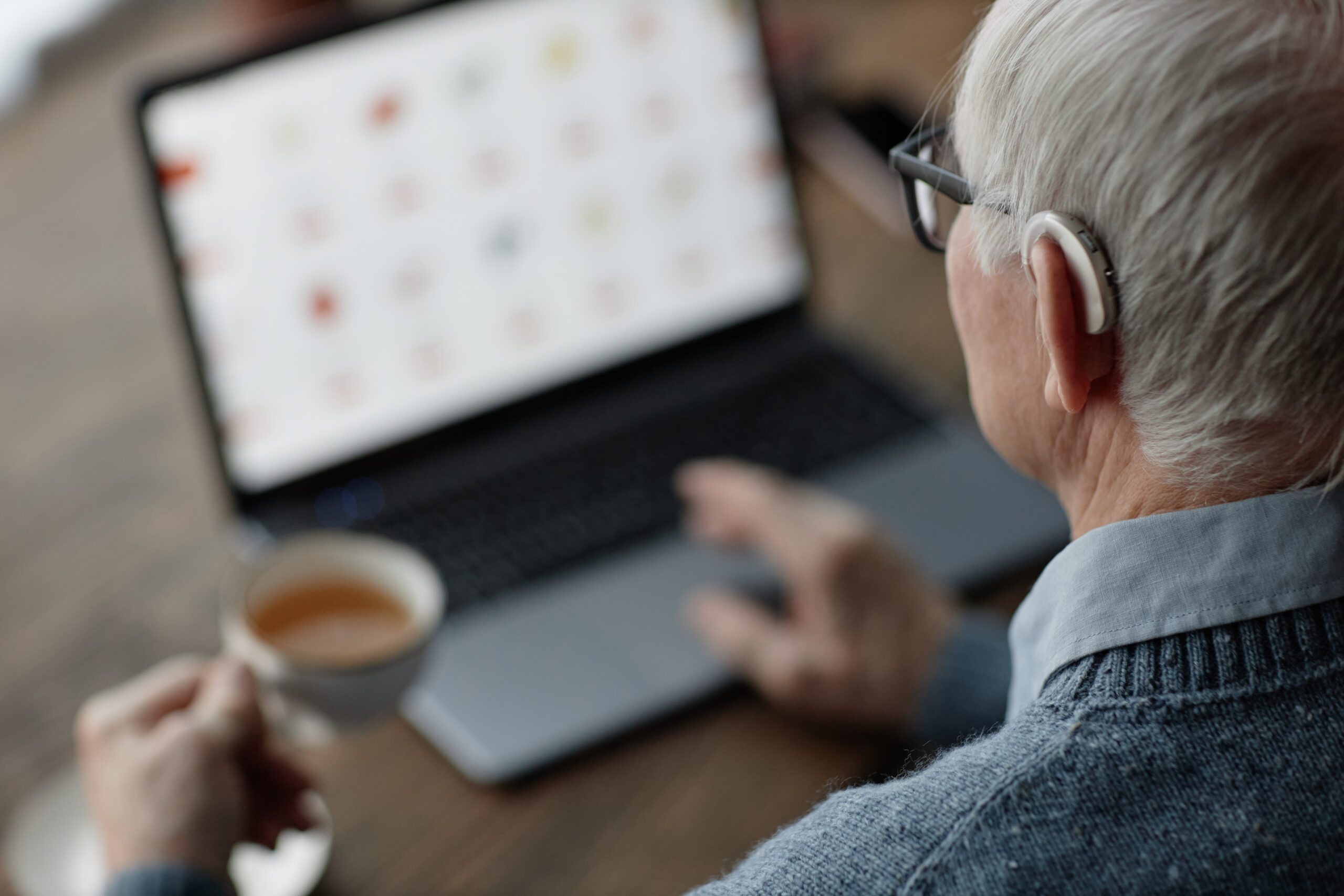 Elderly man wearing hearing aid working on laptop at home while using assistive technology